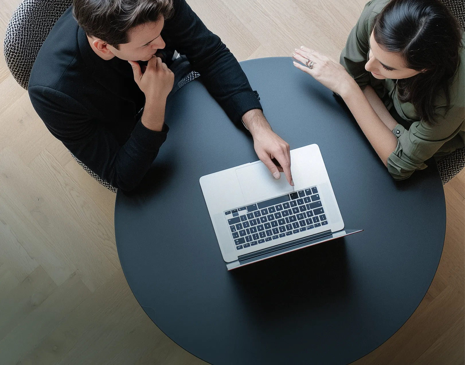 Due persone sedute a un tavolo rotondo nero che guardano uno computer portatile, una indica la tastiera.