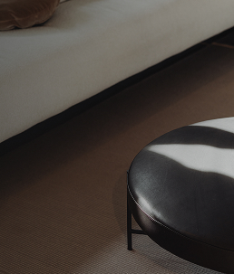 Poggiapiedi rotondo in pelle nera vicino a un divano beige su moquette marrone.