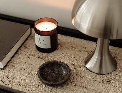 Una candela accesa in un barattolo nero, una ciotola di pietra scura e una lampada in metallo su un tavolo di tessuto beige.