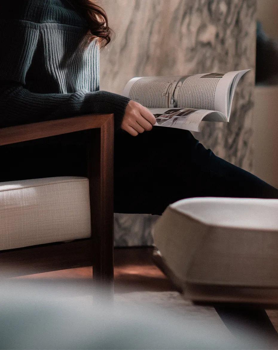 Persona seduta su una poltrona di legno con cuscino beige che tiene in mano una rivista aperta.